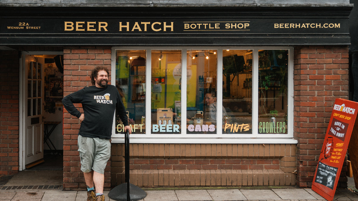 A man wearing a black 'Beer Hatch' t-shirt and khaki shorts stands outside a brick storefront labeled 'BEER HATCH BOTTLE SHOP' with large windows and a sidewalk sign on a paved street.